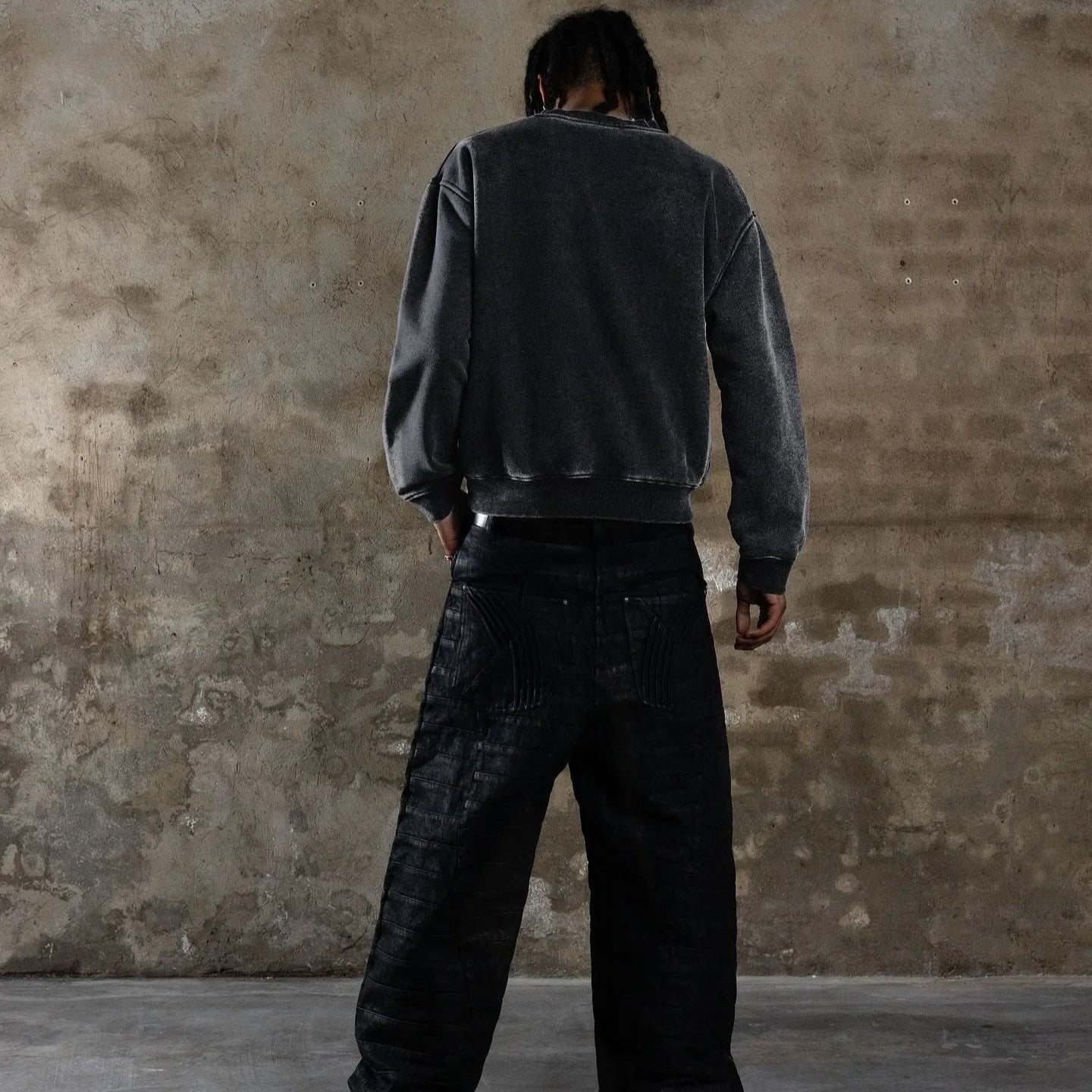 Person wearing a dark gray sweatshirt and black pants standing against a textured wall.