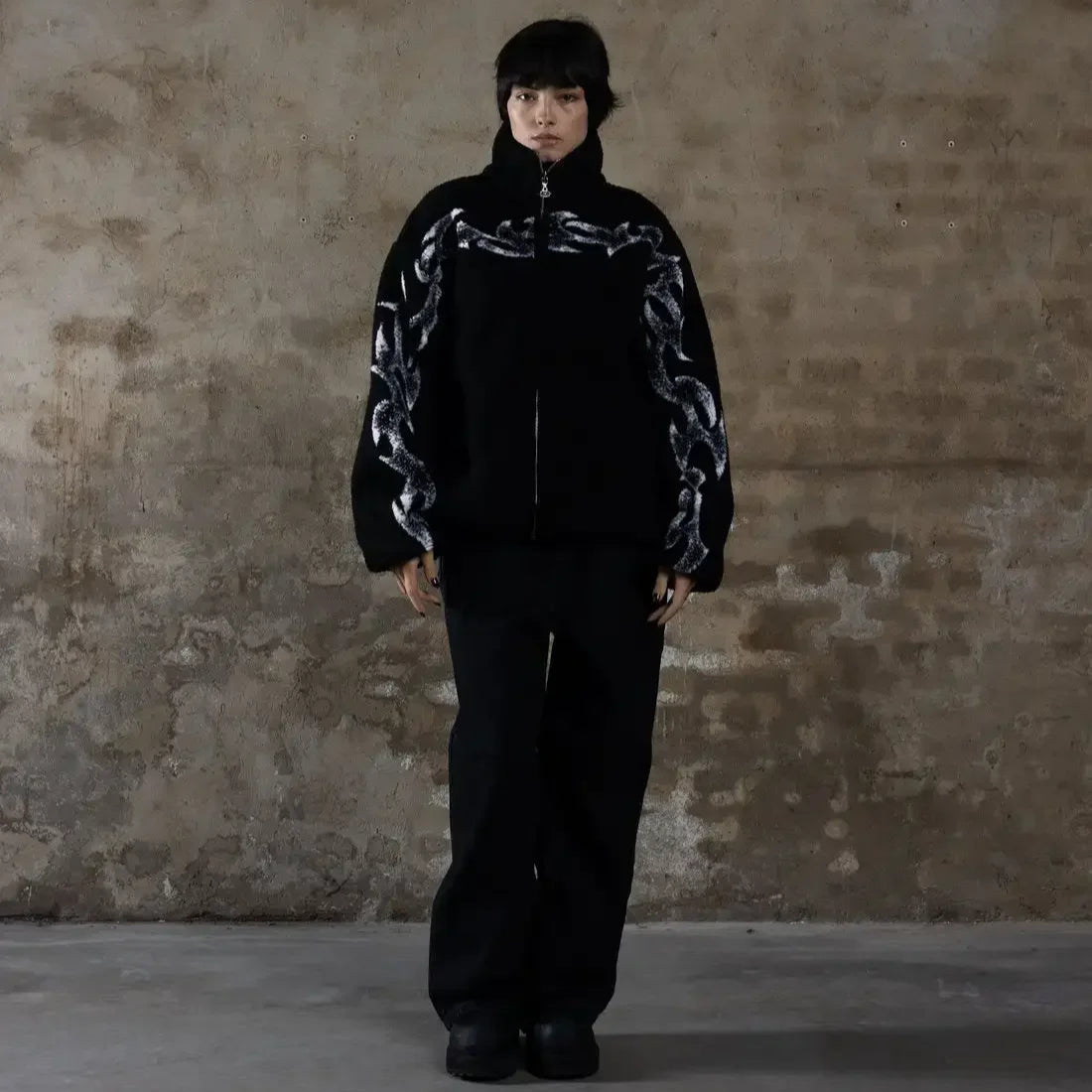 Person wearing a black outfit with patterned sleeves against a textured wall.
