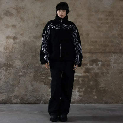 Person wearing a black outfit with patterned sleeves against a textured wall.