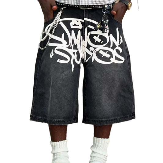 Person wearing black denim shorts with white graphics, white socks, and black shoes against a brick wall.